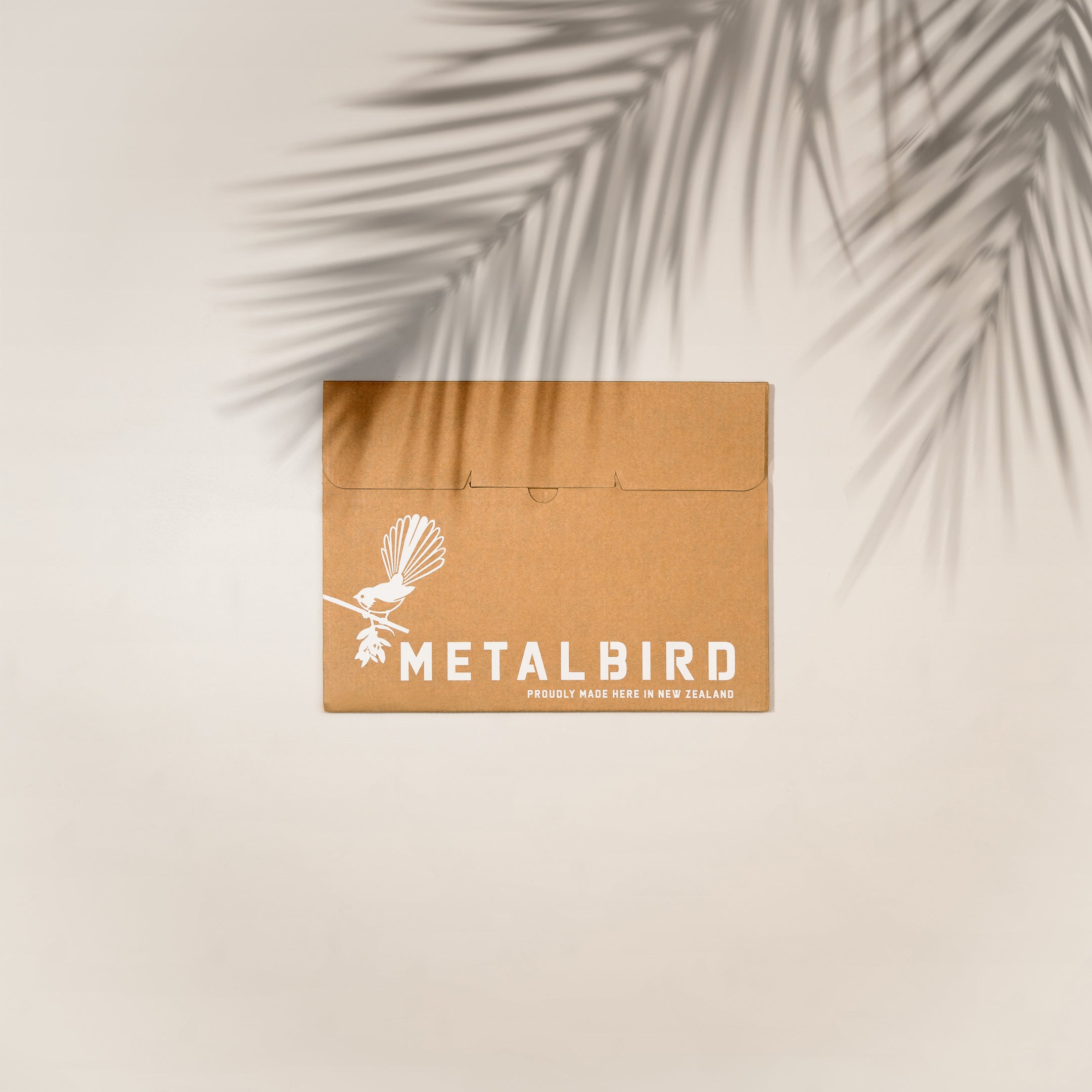 A brown cardboard envelope with the Metalbird New Zealand logo rests on a light background with palm leaf shadows. Text reads: Proudly made here in New Zealand—premium corten steel Hoiho / Yellow-eyed Penguin yard art inside.