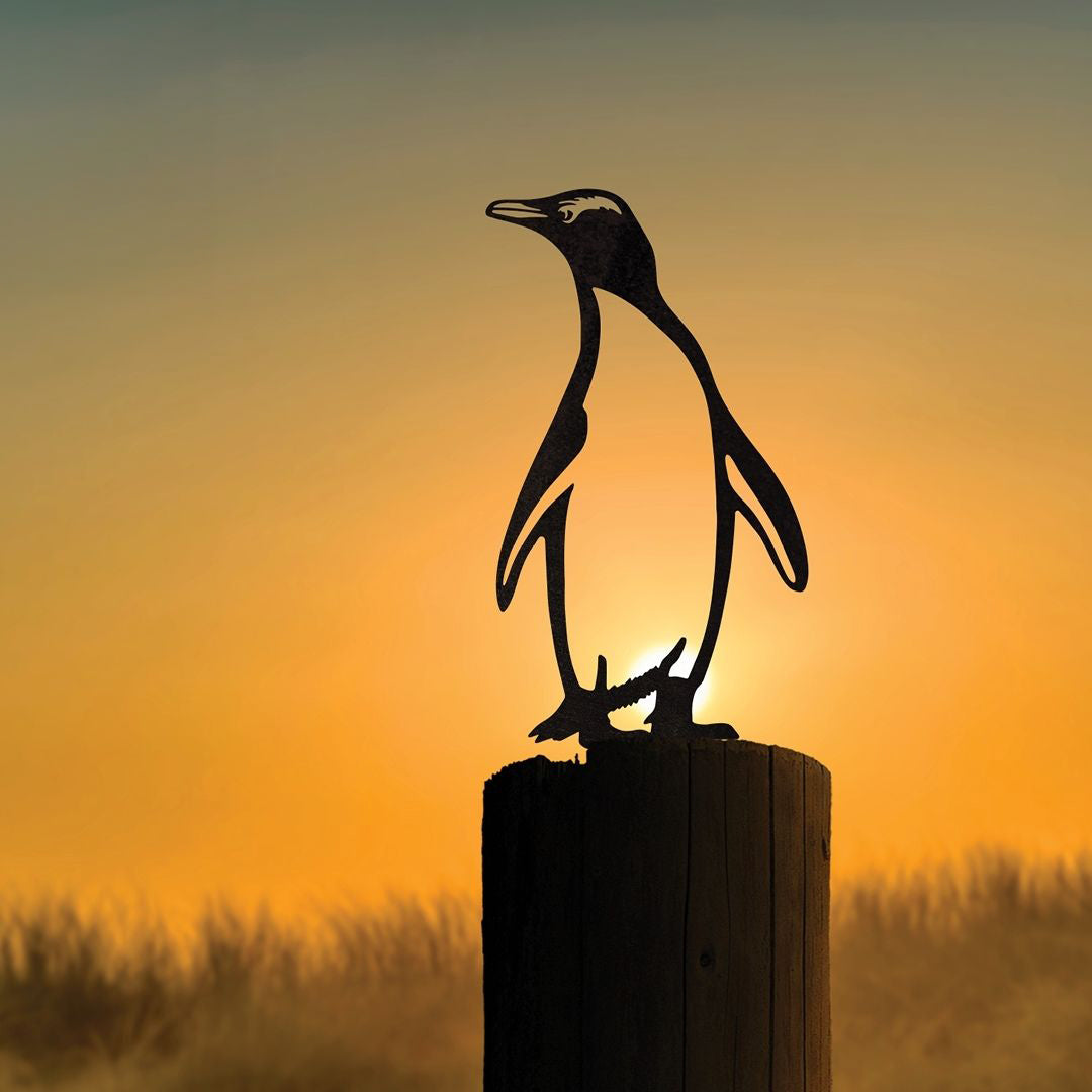 The Metalbird New Zealand Hoiho / Yellow-eyed Penguin is a Corten steel garden art silhouette on a wooden post, shown against a warm orange sunset and blurred grass, celebrating the Bird of the Year.