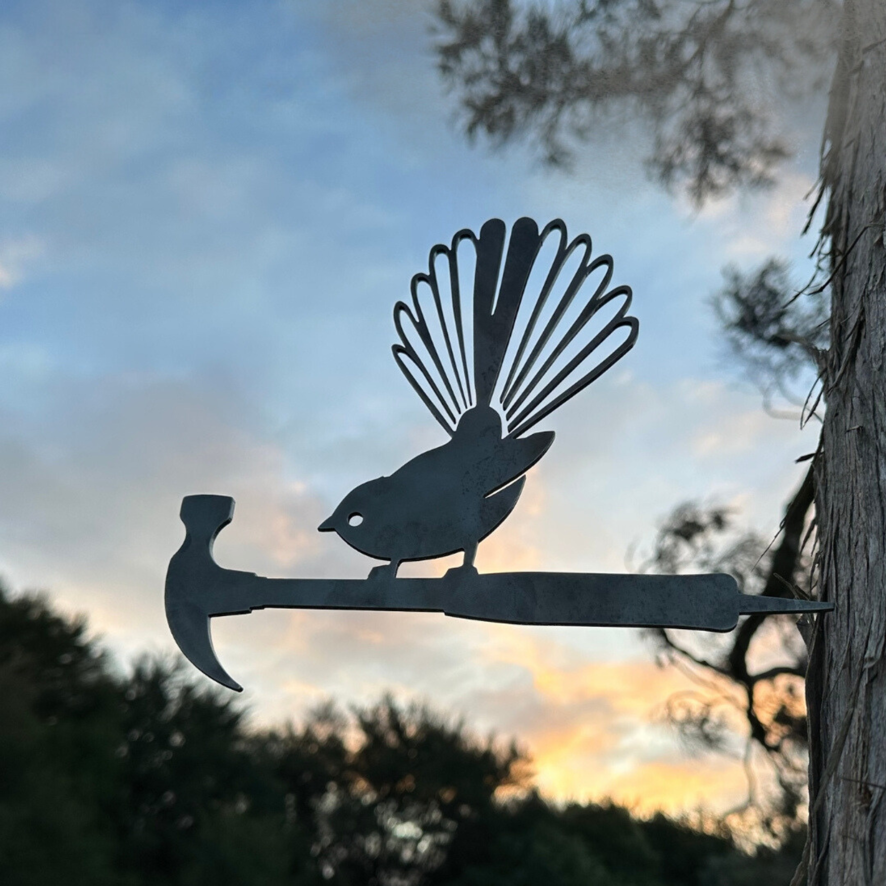 A Fan of D.I.Y. by Metalbird New Zealand—a premium metal bird with a fanned tail perched on a hammer—makes stunning yard art, standing out beautifully against trees and sunset skies.