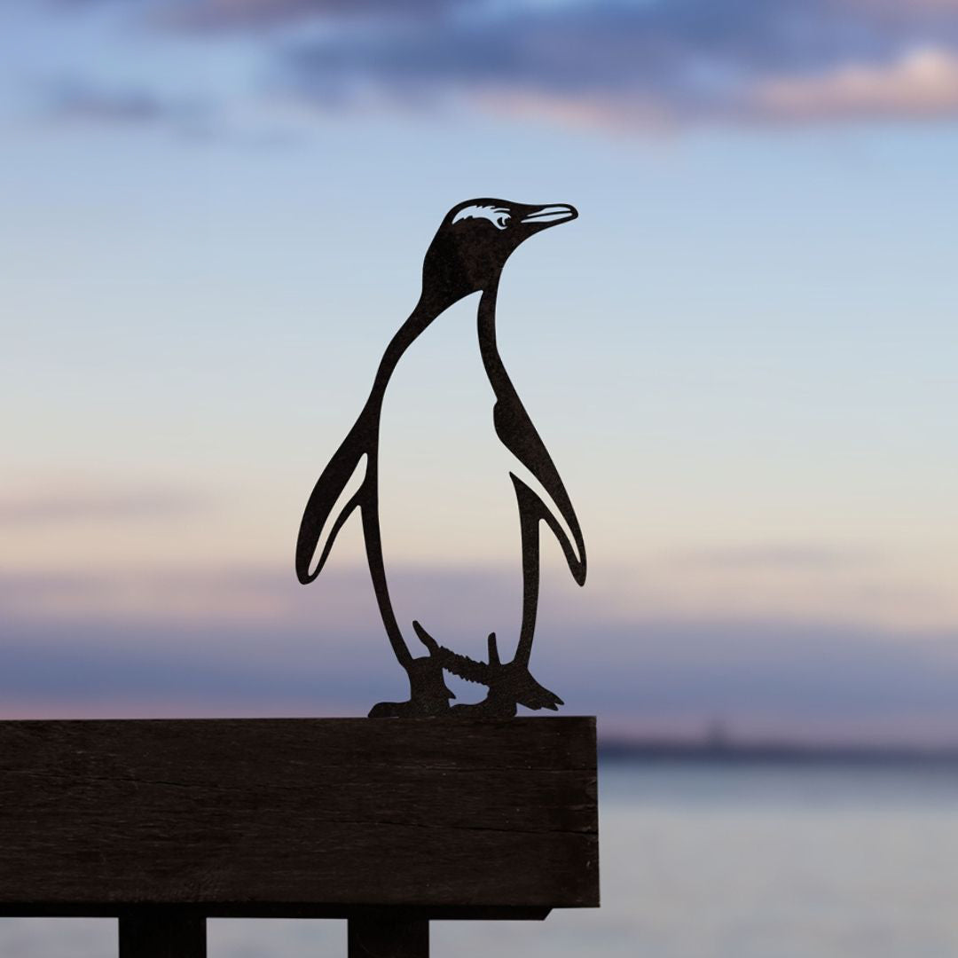 The Metalbird New Zealand Hoiho / Yellow-eyed Penguin Corten steel garden art sculpture stands on a wooden ledge, set against a blurred backdrop of tranquil water and a pastel-hued sunset sky.