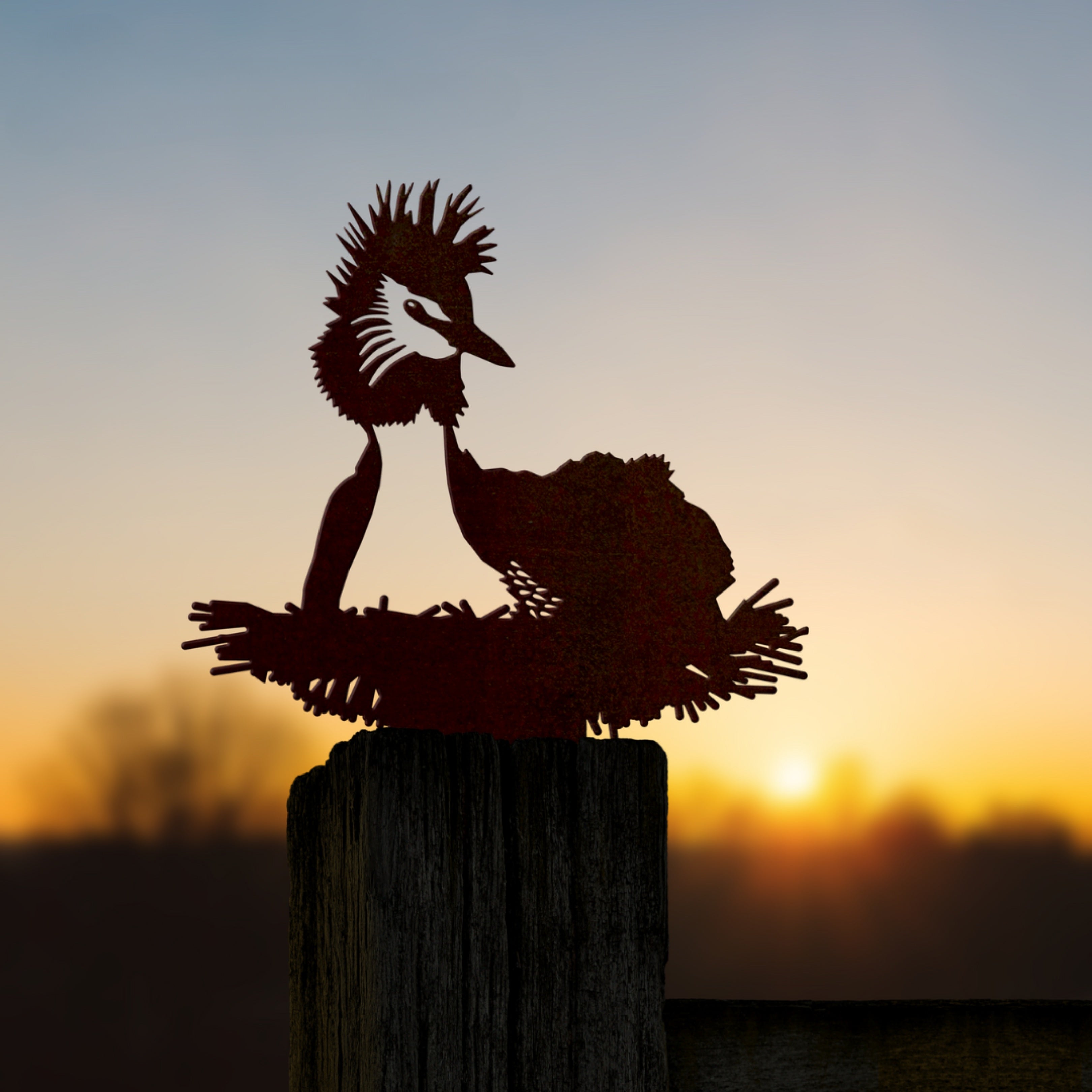 A premium Pūteketeke / Crested Grebe metal silhouette by Metalbird New Zealand is mounted on a wooden post against a soft-focus sunset and trees—an ideal gift for nature lovers.