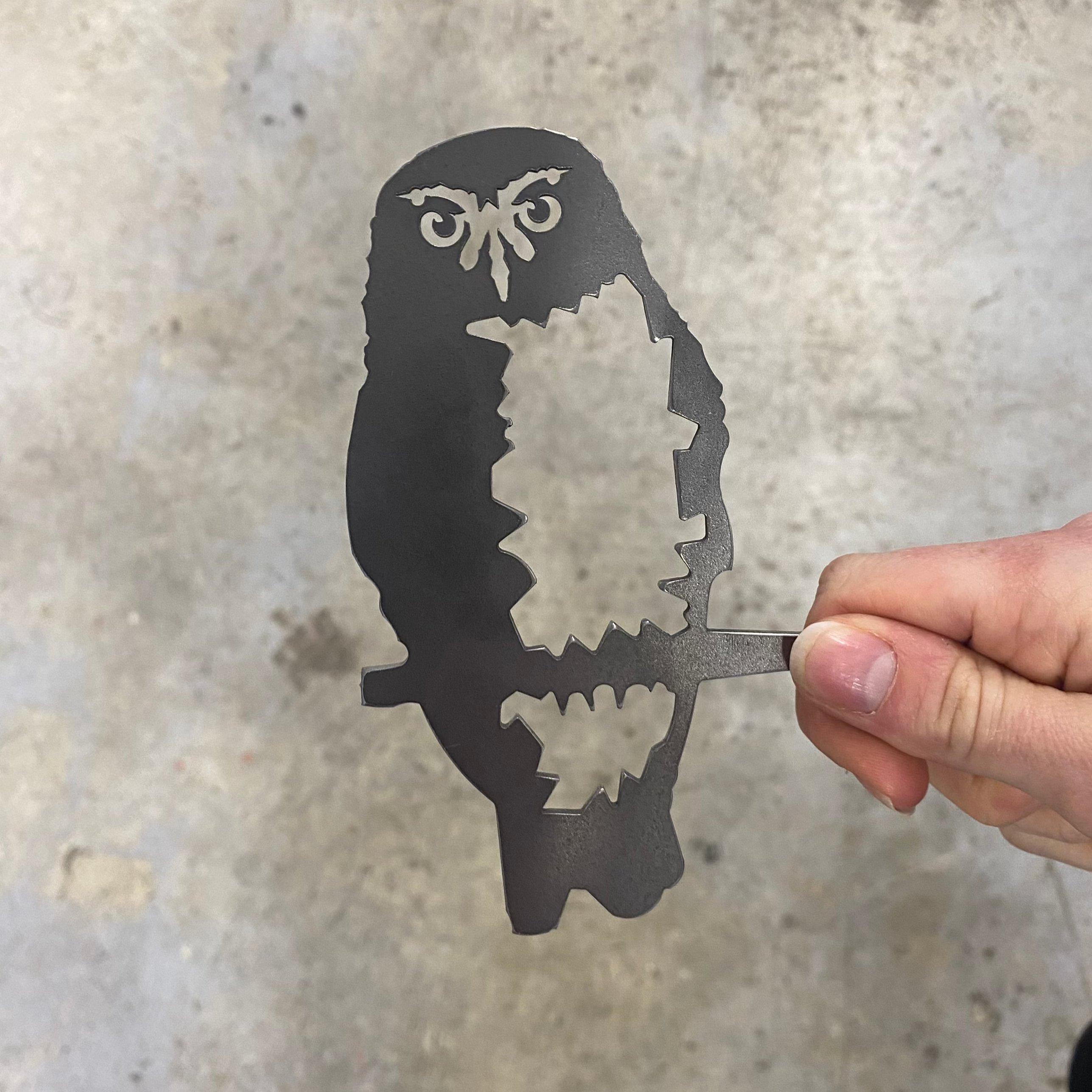 A hand holds the Metalbird New Zealand Mini Ruru, a premium corten steel art cutout shaped like an owl on a branch, featuring detailed metalwork for the eyes, beak, and body, set against a concrete background.