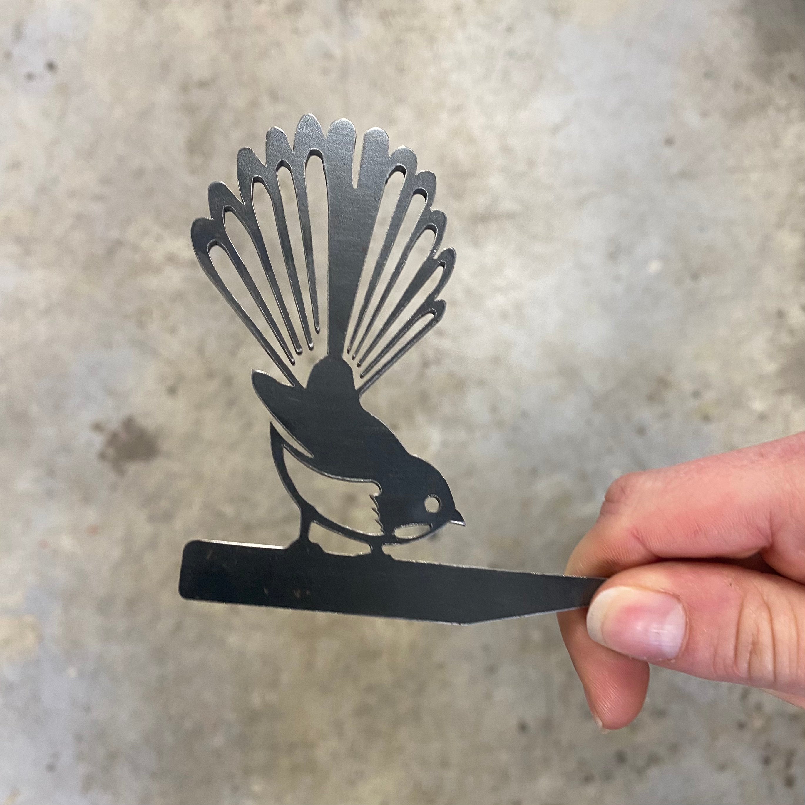 A hand holds the Mini Pīwakawaka, a black corten steel bird silhouette by Metalbird New Zealand, its fanned tail resembling a fantail, set against a light textured background.