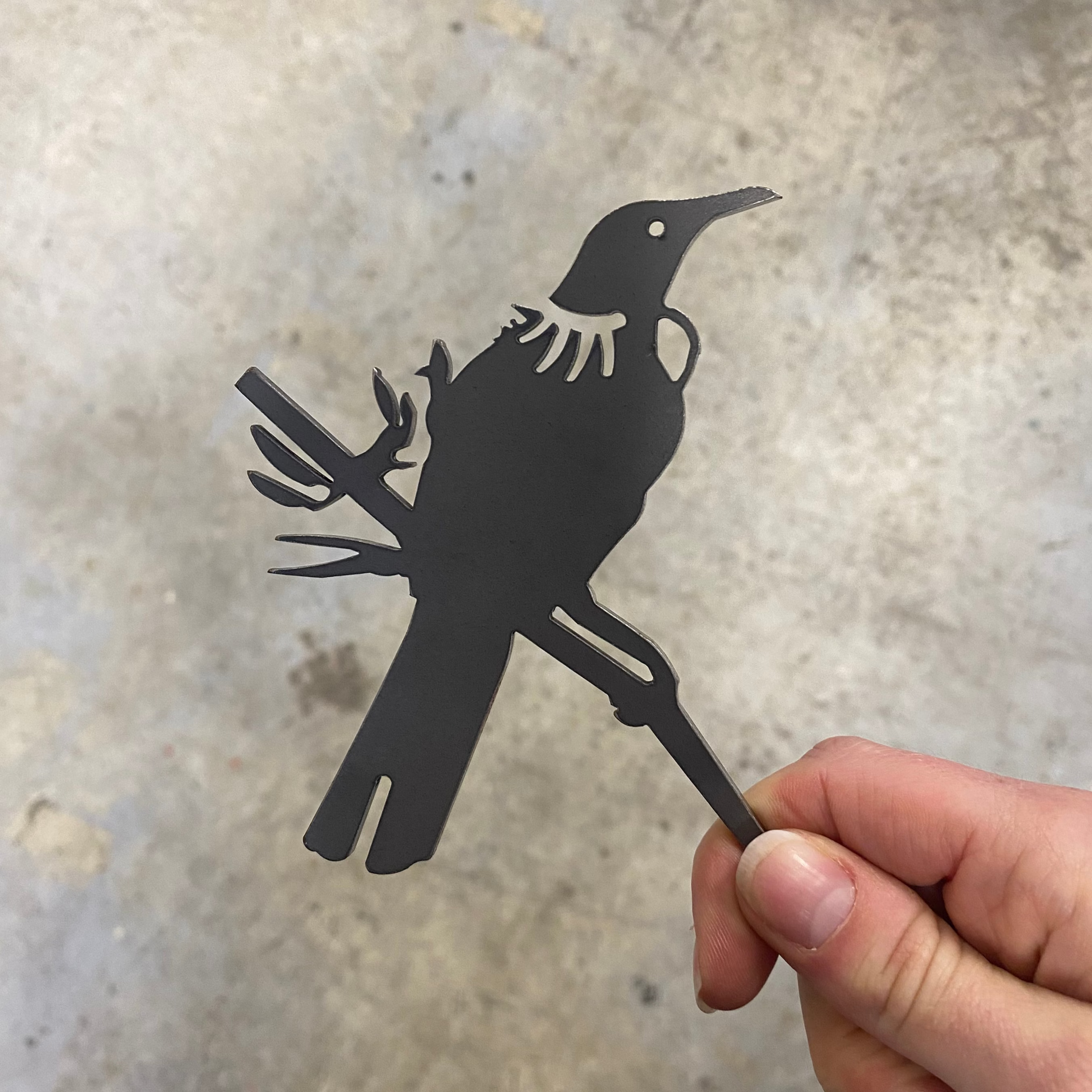 A hand holds the Mini Tūī by Metalbird New Zealand, a flat black corten steel bird silhouette, perfect as unique yard art, seen against a blurred gray background.