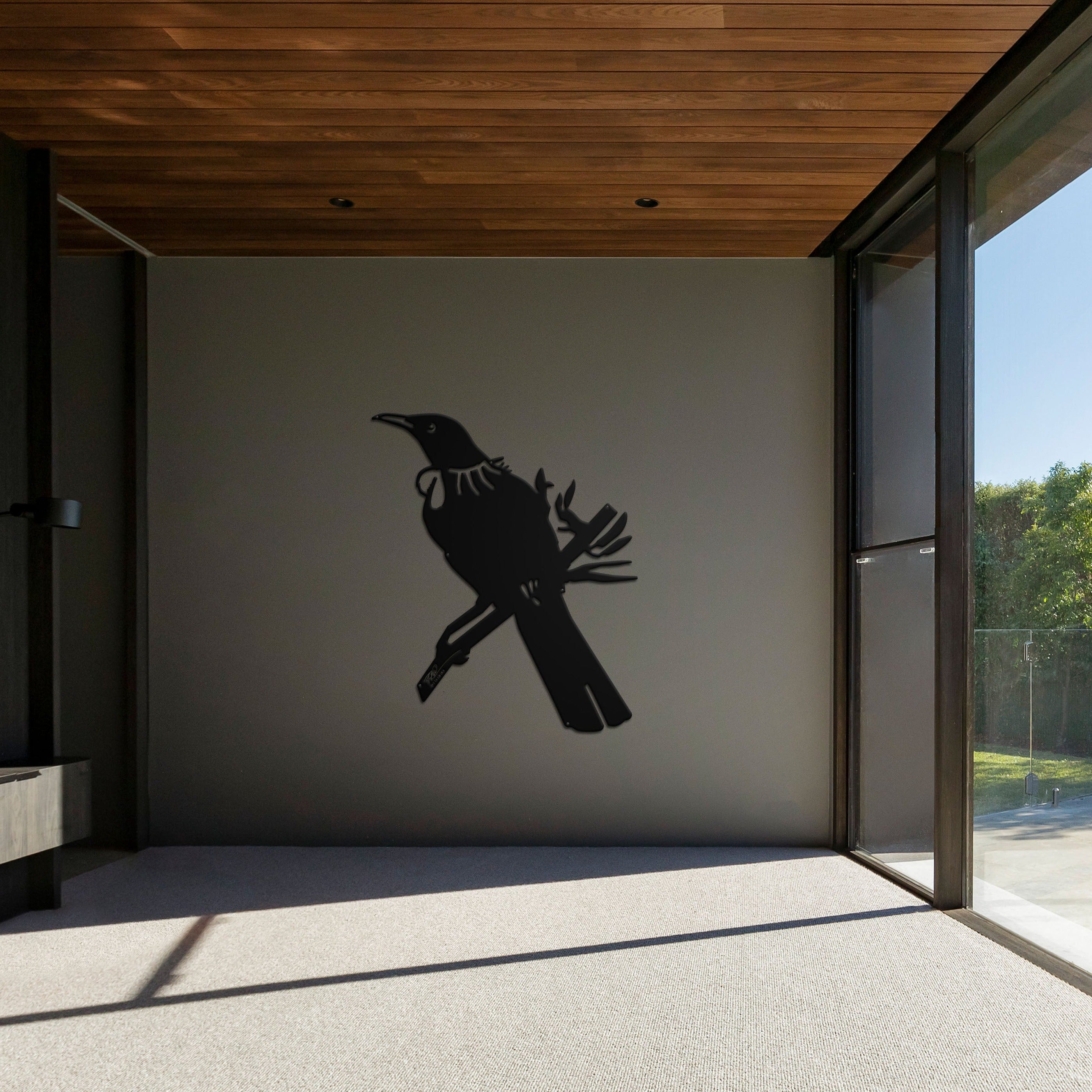 A modern, sunlit room with large windows showcases the Tūī Big Bird Wall Sculpture by Metalbird New Zealand—a striking silhouette of a bird on a plain gray wall. The wood-paneled ceiling and trees outside add warmth and serenity.