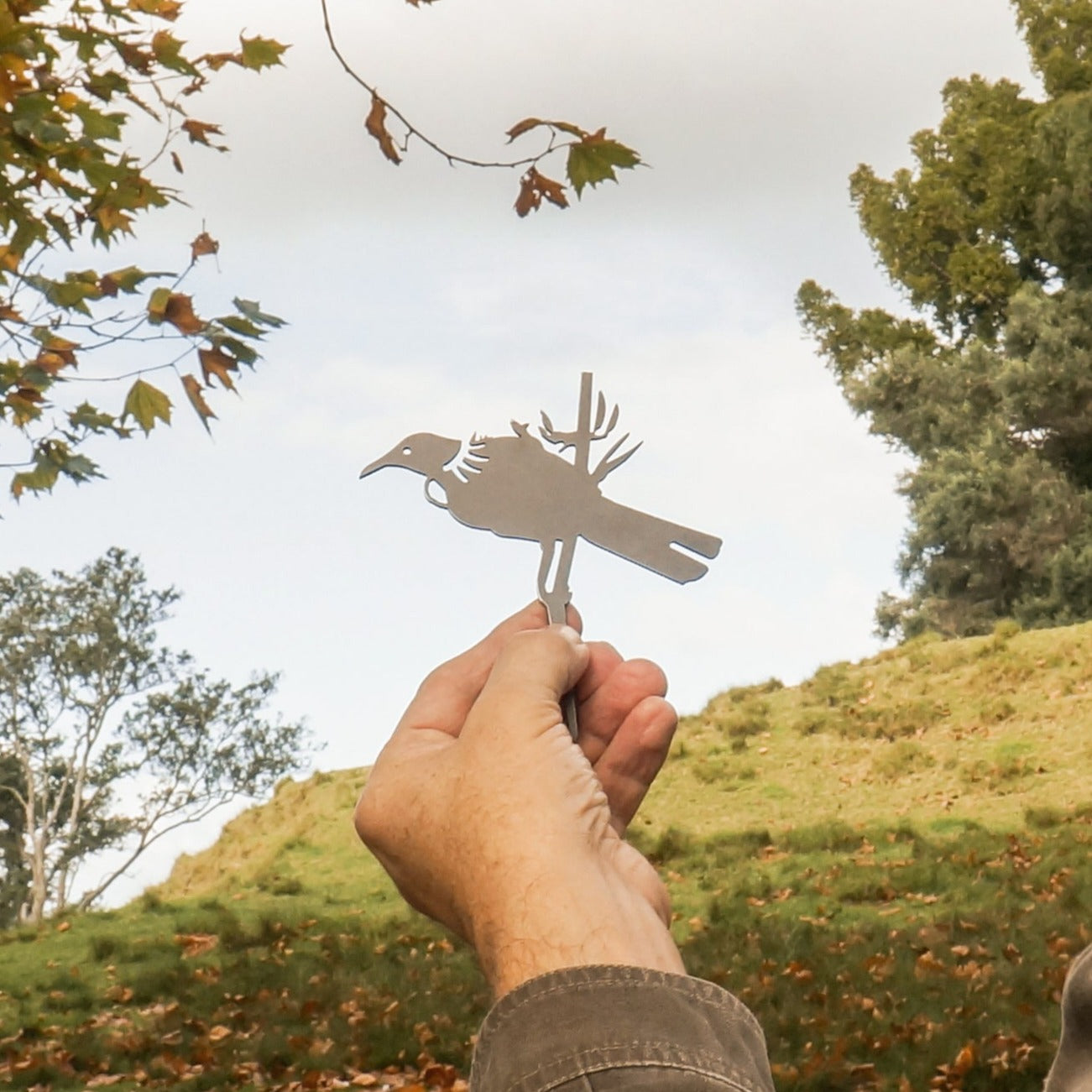 A hand holds up the Mini Tūī metal silhouette by Metalbird New Zealand, featuring a bird with a branch design, set against an outdoor backdrop of trees, grassy hillside, and a partly cloudy sky.