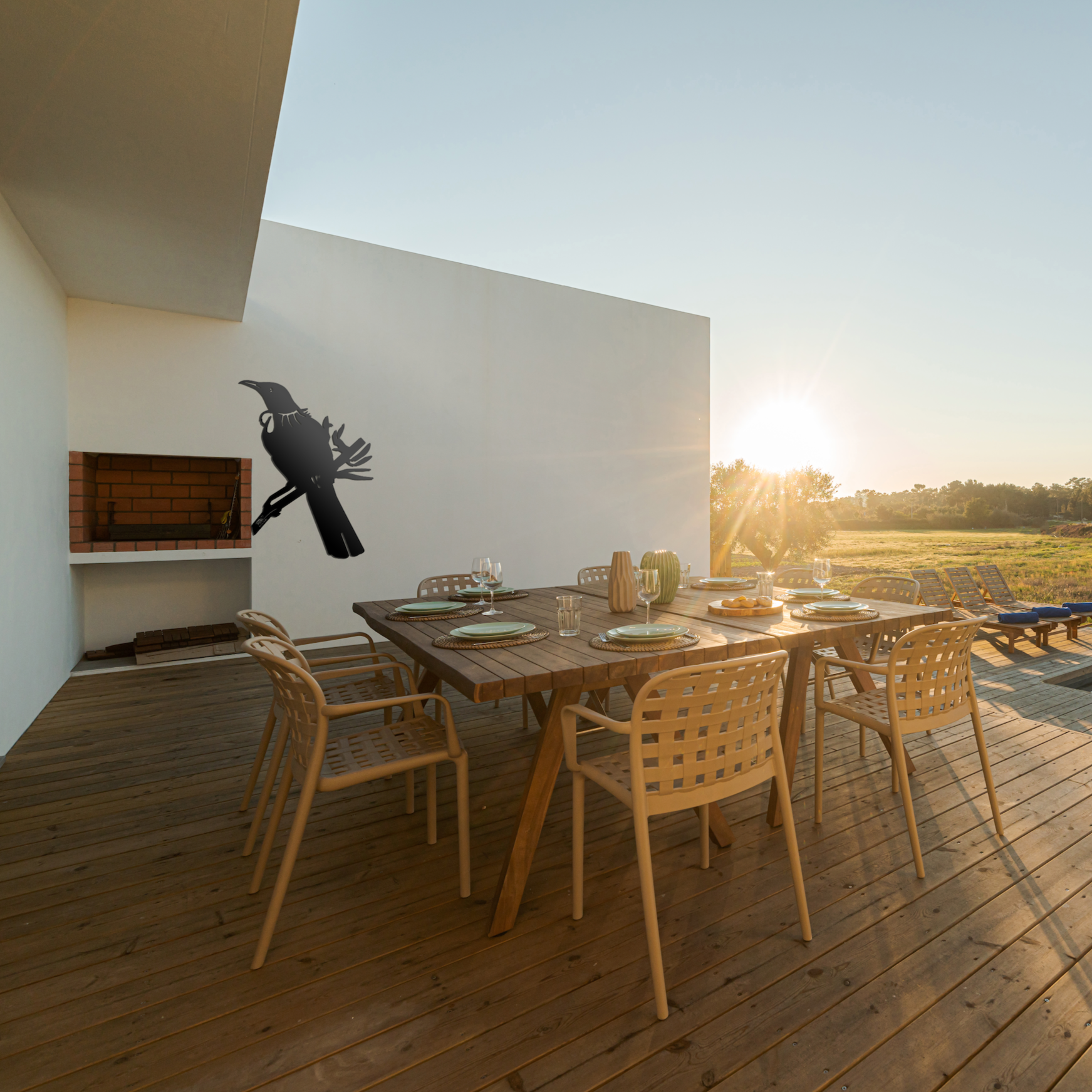 Modern outdoor dining area at sunset with plates and glasses on a wooden deck. A minimalist barbecue is built into a white wall adorned with the Tūī Big Bird Wall Sculpture by Metalbird New Zealand, adding striking art to the yard.