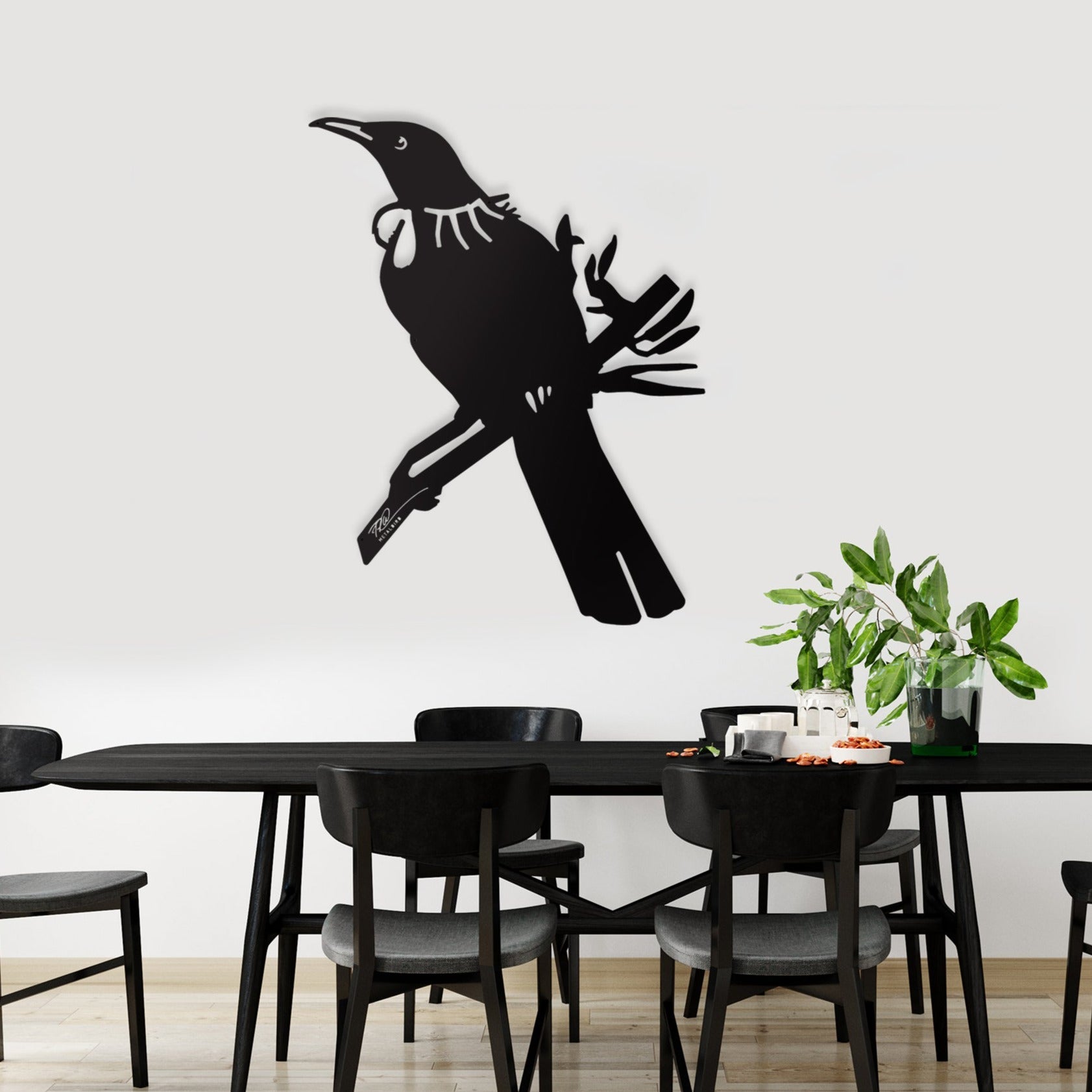 A modern dining room features a black table and chairs, a plant centerpiece, and the Metalbird New Zealand Tūī Big Bird Wall Sculpture—a striking corten steel silhouette of a tūī perched on a branch—an ideal gift for any contemporary space.