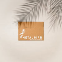 A brown cardboard envelope with the Metalbird New Zealand logo and bird art sits on a light background with palm leaf shadow. Text says: Proudly made in NZ—the perfect packaging for your Quail Mum & Mini Set garden gift.