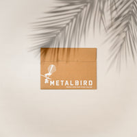 A brown cardboard envelope printed with “Metalbird” and a bird graphic sits on a light backdrop with palm leaf shadows—perfect packaging for the Tūī Mum & Mini Set by Metalbird New Zealand, an ideal Mother's Day gift.