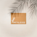A brown cardboard envelope with METALBIRD and a bird graphic in white rests on a light background with palm leaf shadows—an ideal Mother's Day gift: the Pīwakawaka Mum & Mini Set from Metalbird New Zealand.