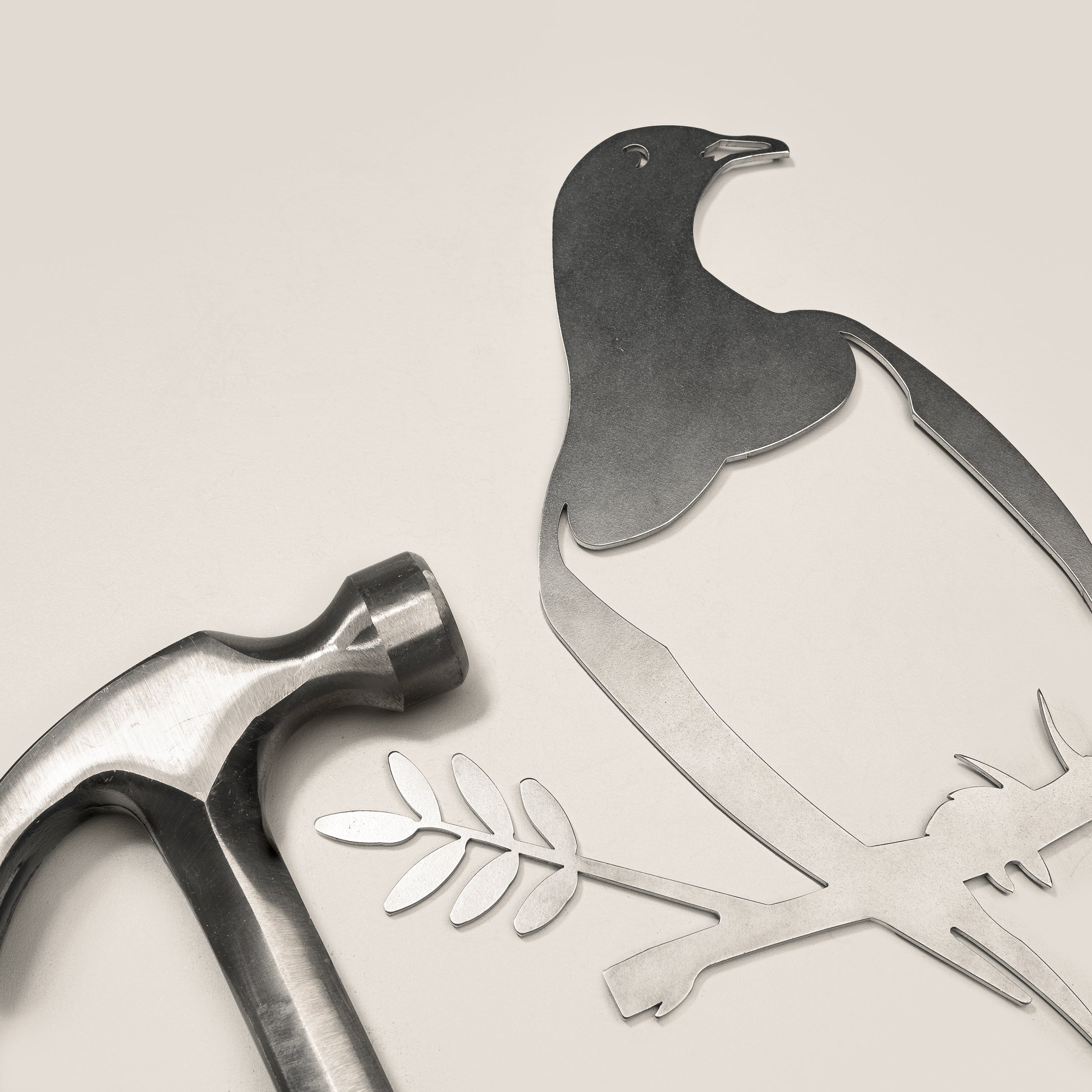 A metal hammer lies beside the Metalbird New Zealand Kererū Mum & Mini Set—a flat metal cut-out of a bird and baby on a branch with leaves—on a smooth, light surface, making it an ideal Mother's Day gift.