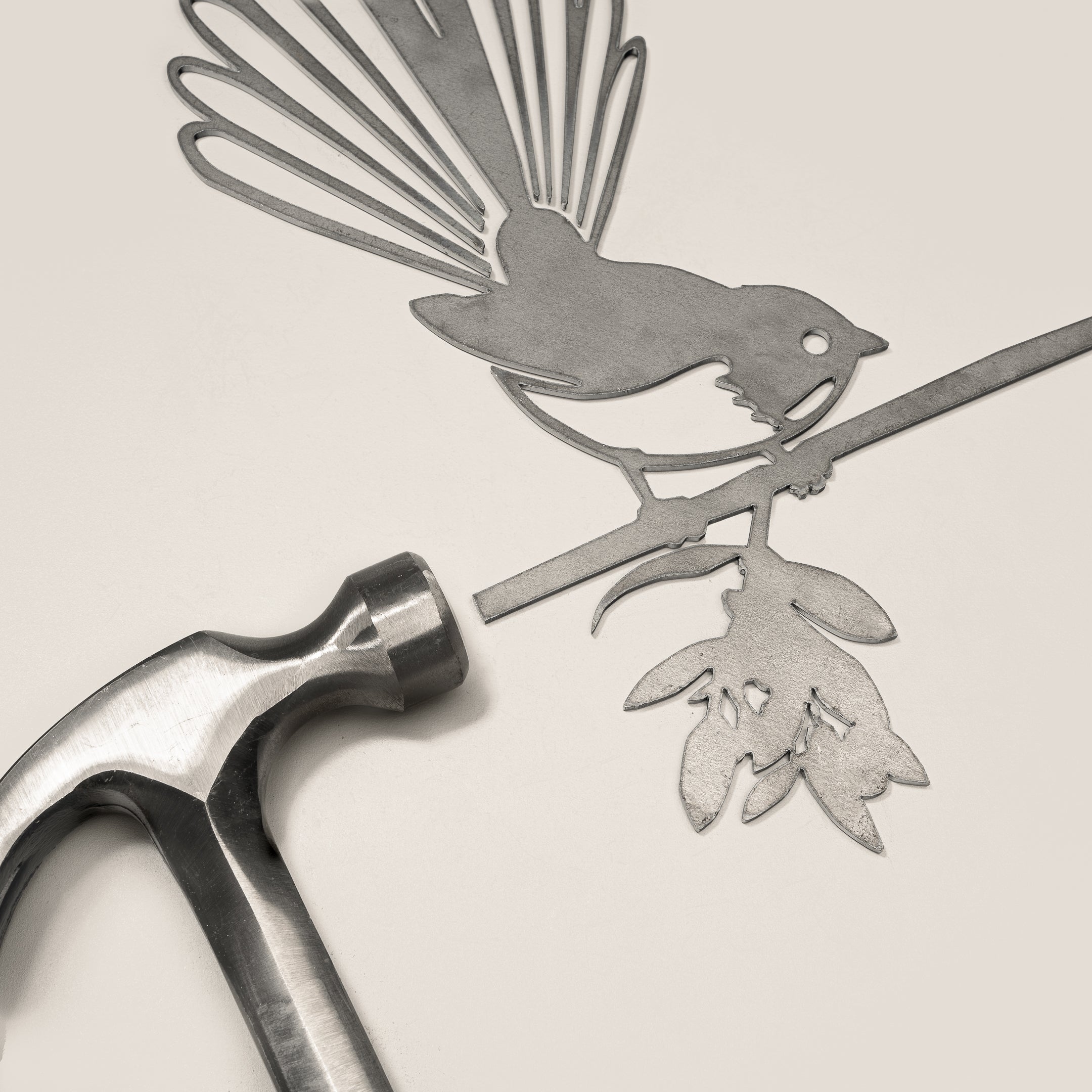 The Pīwakawaka Mum & Mini Set by Metalbird New Zealand features a delicate metal cutout of a Pīwakawaka bird and chick, crafted for a special Mother’s Day gift, displayed next to a metal hammer on a light surface.