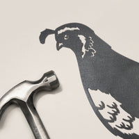 A metal hammer lies next to a flat, life-sized quail silhouette by Metalbird New Zealand—perfect as a garden gift in NZ or to complement your Quail Mum & Mini Set.