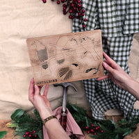 A person holds a wooden board displaying Winged Hangers - 5 Festive Decorations by Metalbird New Zealand. Holiday greenery, a hammer, fabric, and red berries set a festive crafting scene with sustainably made in Aotearoa details.