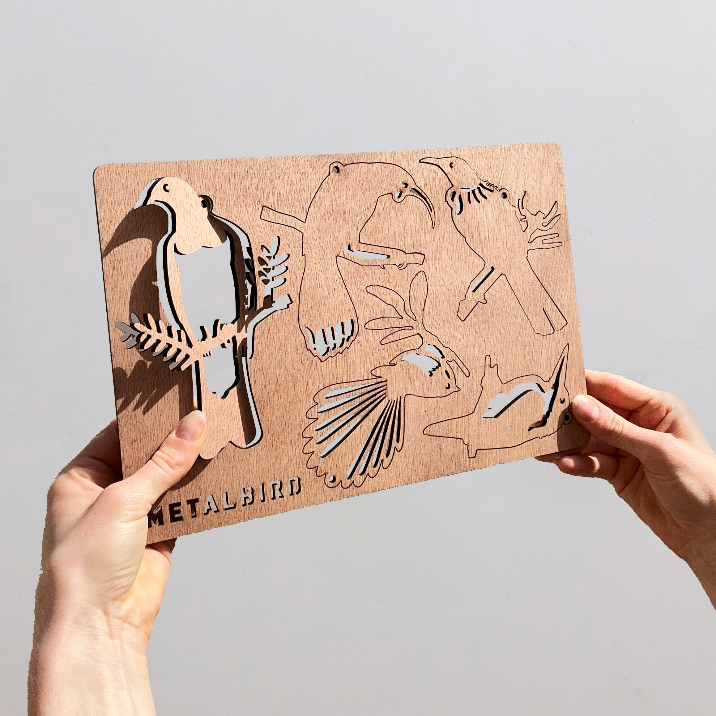 A person holds a brown metal stencil from Metalbird New Zealand, featuring five festive Winged Hangers bird silhouettes in various poses. "METALBIRD" is engraved at the bottom left; the background is plain and light-colored.