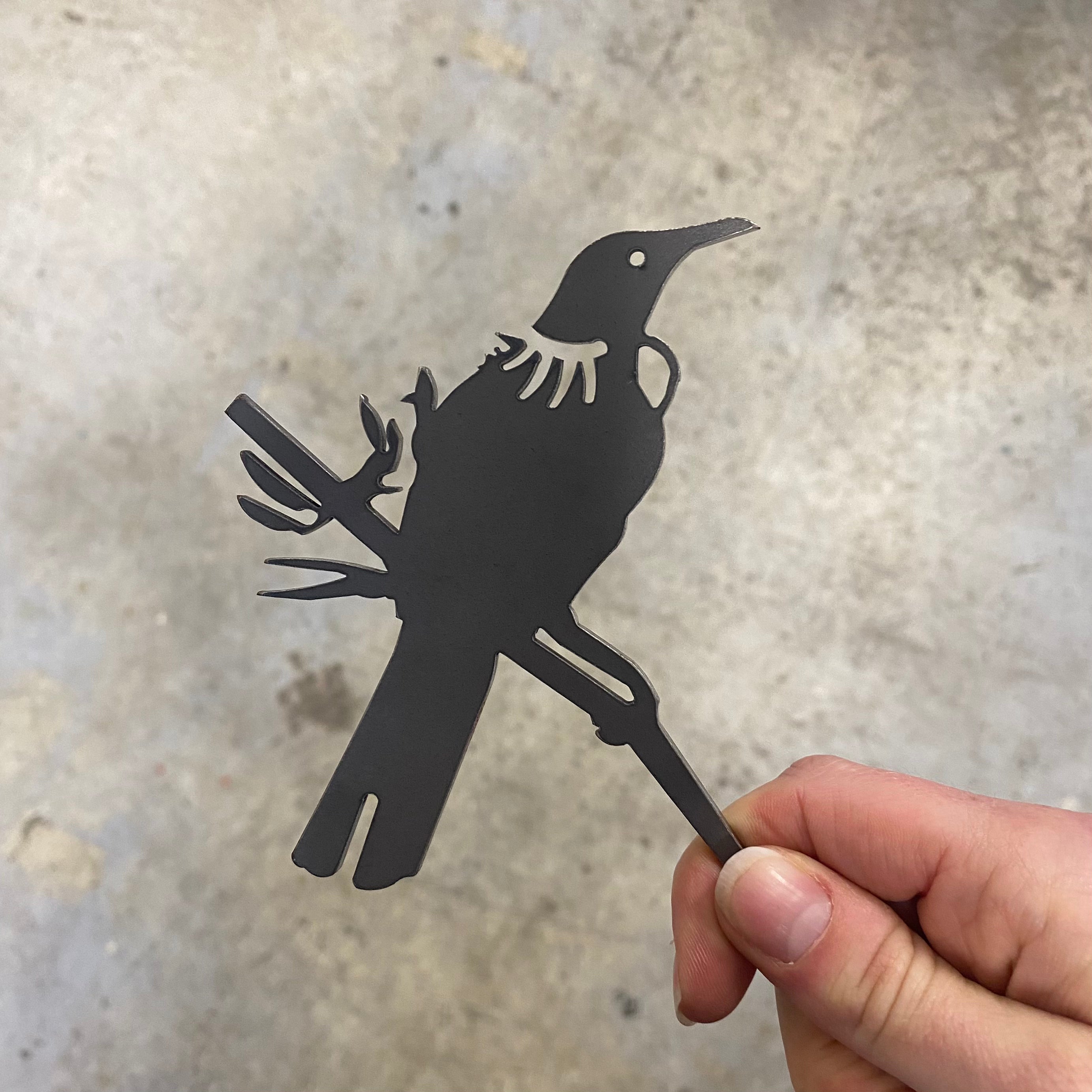 A hand holds the Metalbird New Zealand Tūī Mum & Mini Set, a metal silhouette of a mother and baby Tūī on a branch—perfect as a garden sculpture or Mother's Day gift—set against a blurred gray background.