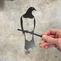 A hand holds the Kererū Mum & Mini Set by Metalbird New Zealand—a flat, black metal silhouette of a mum and baby Kererū on a branch, set against a light gray marbled background. The perfect Mother’s Day gift inspired by an iconic NZ bird.