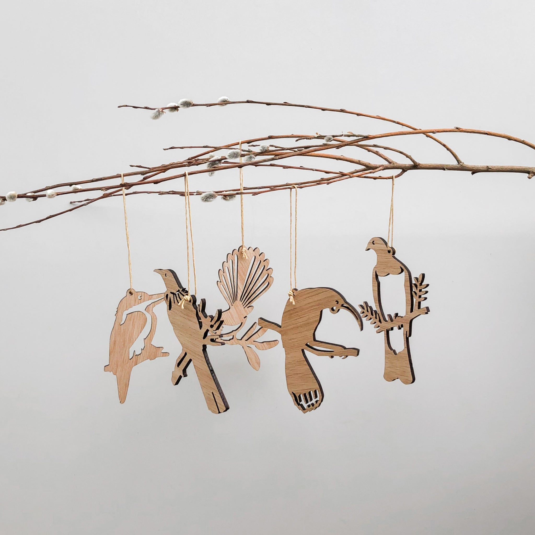 The Winged Hangers 5 Festive Decorations by Metalbird New Zealand feature bird and shell silhouettes hanging from strings on a bare branch, sustainably crafted in Aotearoa for an eco-friendly touch against a plain, light background.