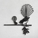 The metalbird Metalbird Seconds - PIWAKAWAKA AND BABY - XL features two stylized birds with fan-shaped tails, perched on a branch; its metal surface has imperfections and looks striking against a textured white wall.