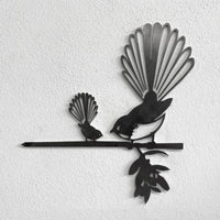 The metalbird Metalbird Seconds - PIWAKAWAKA AND BABY - XL features two stylized birds with fan-shaped tails, perched on a branch; its metal surface has imperfections and looks striking against a textured white wall.