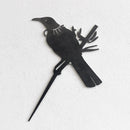 A black metal silhouette of a piwakawaka and baby perched on a branch, set against a white textured wall. This XL stylized cutout from metalbird is part of our Metalbird Seconds range and may feature minor imperfections.