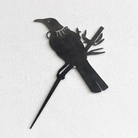 A black metal silhouette of a piwakawaka and baby perched on a branch, set against a white textured wall. This XL stylized cutout from metalbird is part of our Metalbird Seconds range and may feature minor imperfections.