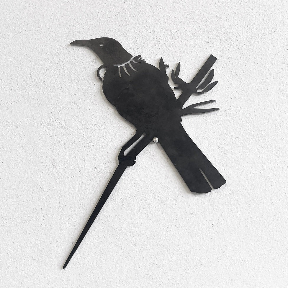 A black metal silhouette of a piwakawaka and baby perched on a branch, set against a white textured wall. This XL stylized cutout from metalbird is part of our Metalbird Seconds range and may feature minor imperfections.