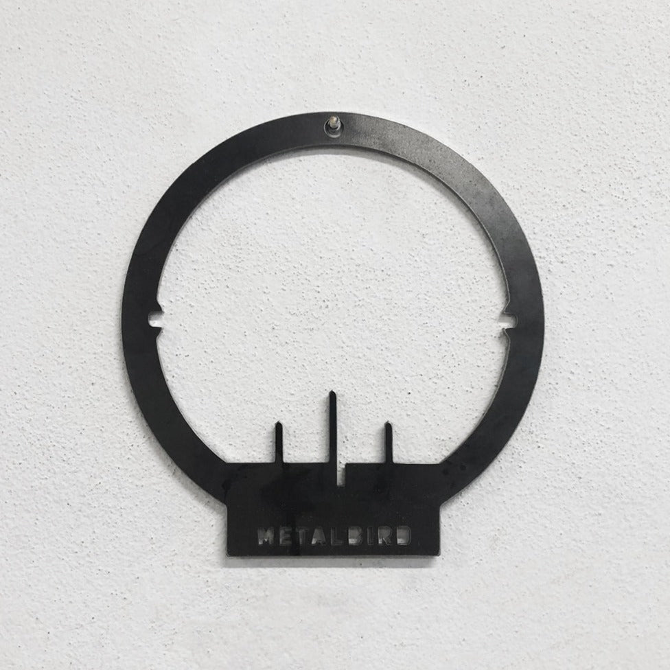 Mounted on a textured white wall, the Metalbird Seconds - PIWAKAWAKA AND BABY - XL by metalbird is a unique black metal circular wall decor featuring three vertical bars and METALBIRD cutout at the base, each with its own imperfections.