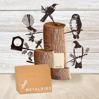 A display of Metalbird New Zealand bird silhouettes on wooden logs with envelopes and the Christmas Favourites Gift Pack, all set against light wood paneling.