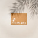 A brown cardboard envelope with Metalbird New Zealand and a bird graphic in white, perfect for gifting the Pūkeko sculpture, rests on a light surface with a palm leaf shadow across the top right corner.