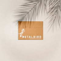 A brown cardboard envelope with Metalbird New Zealand and a bird graphic in white, perfect for gifting the Pūkeko sculpture, rests on a light surface with a palm leaf shadow across the top right corner.