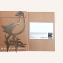 A Kiwiana gift by Metalbird New Zealand features a corten steel Pūkeko bird with grass, attached to an open brown folder holding a white info card and logos, including Forest & Bird, displayed on the right.