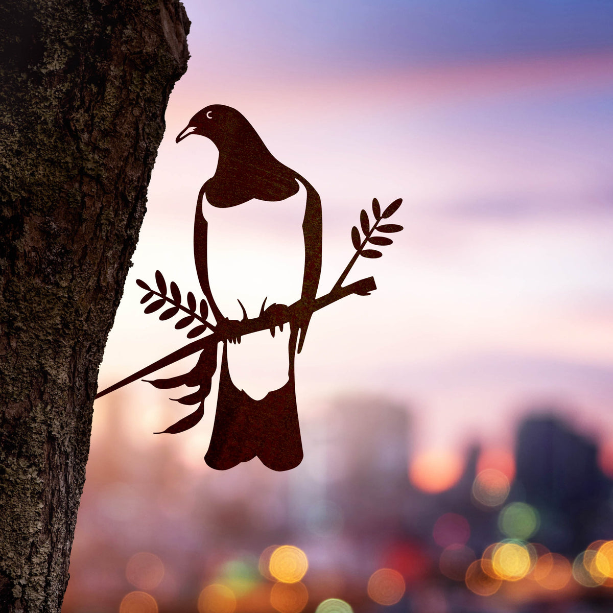 The Kererū / Wood Pigeon by Metalbird New Zealand is a premium corten steel silhouette of a bird with an olive branch, mounted on a tree trunk—set against a sunset cityscape—making it the perfect gift.