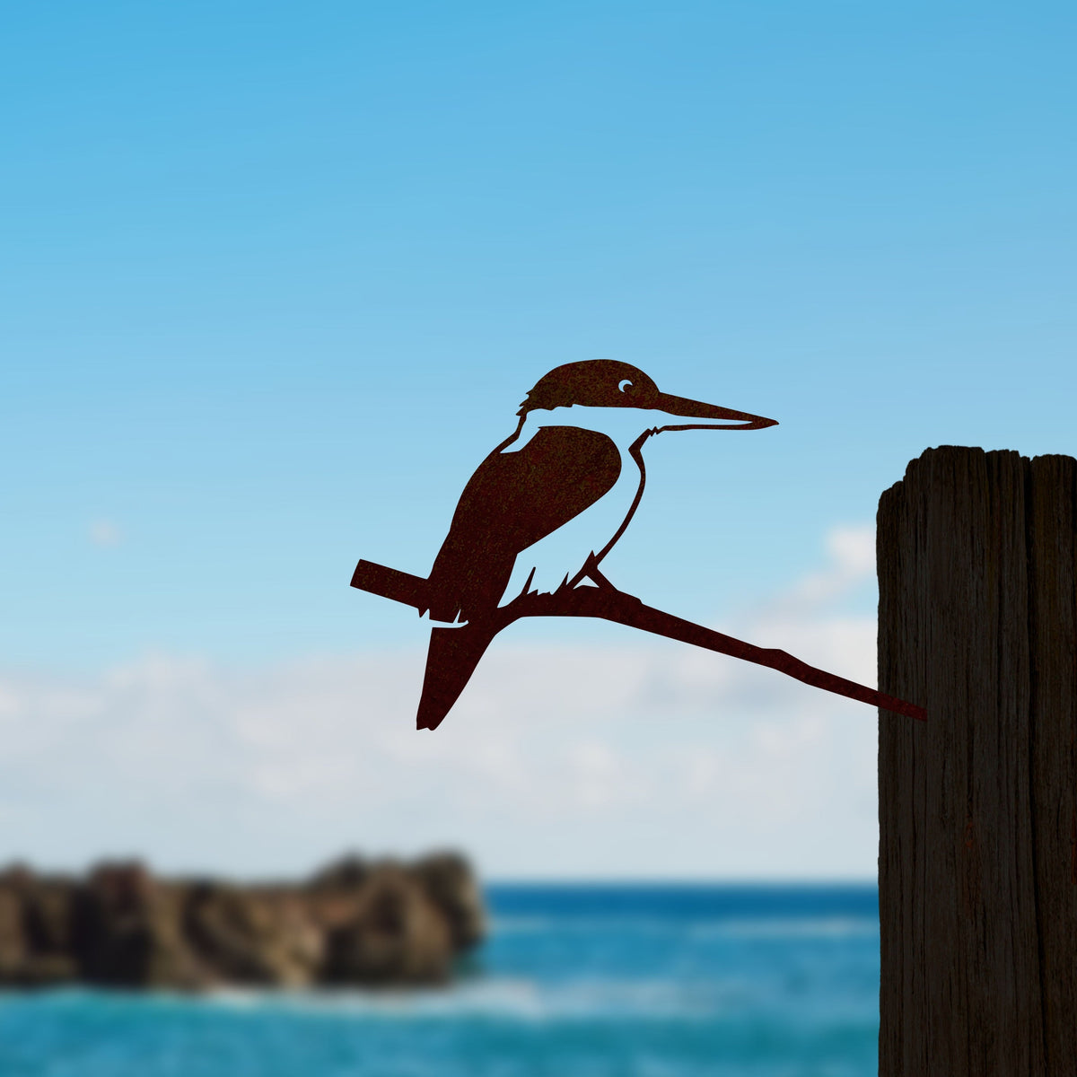 Kōtare steel silhouette mounted beside a timber post with sea and rocks behind.