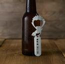 A brown glass bottle sits on wood with the Metalbird New Zealand Birdle-Opener 2.0, a stainless steel, bird-shaped opener engraved with METALBIRD, leaning against it.