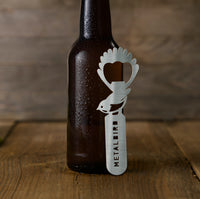 A brown glass bottle sits on wood with the Metalbird New Zealand Birdle-Opener 2.0, a stainless steel, bird-shaped opener engraved with METALBIRD, leaning against it.