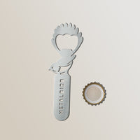 The Metalbird New Zealand Birdle-Opener 2.0, a stainless steel bird-shaped bottle opener with METALBIRD cut into the handle, rests beside a removed cap on a light background.