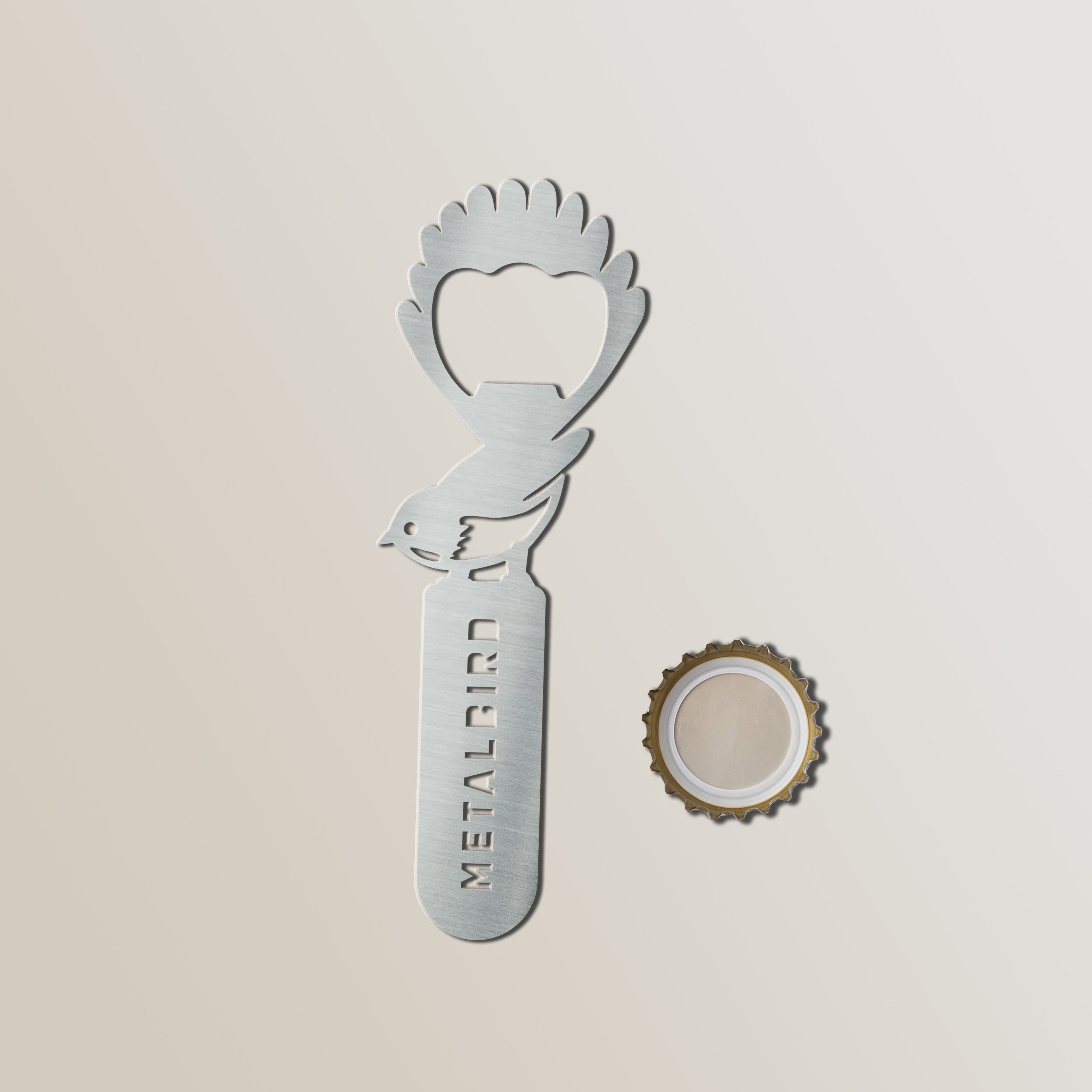 The Metalbird New Zealand Birdle-Opener 2.0, a stainless steel bird-shaped bottle opener with METALBIRD cut into the handle, rests beside a removed cap on a light background.