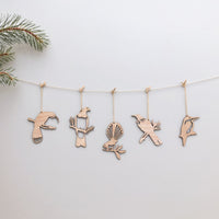 Five sustainably made Metalbird New Zealand Winged Hangers—each shaped like a different bird—hang from a string with clothespins. A pine branch accents the corner, set against a plain white backdrop. Set includes 5 festive decorations.