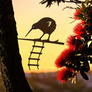 The KIWI : Glenn Jones Art Collaboration by Metalbird New Zealand features a metal kiwi bird on a branch with a hanging ladder, set against a yellow sunset with red flowers and leaves—classic Kiwiana-inspired art.