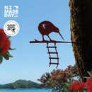 The “KIWI : Glenn Jones Art Collaboration” by Metalbird New Zealand features a metal kiwi bird on a branch with a ladder, framed by pohutukawa blooms, blue sky, and ocean. Includes NZ Made Day and Glenn Jones Art logos.
