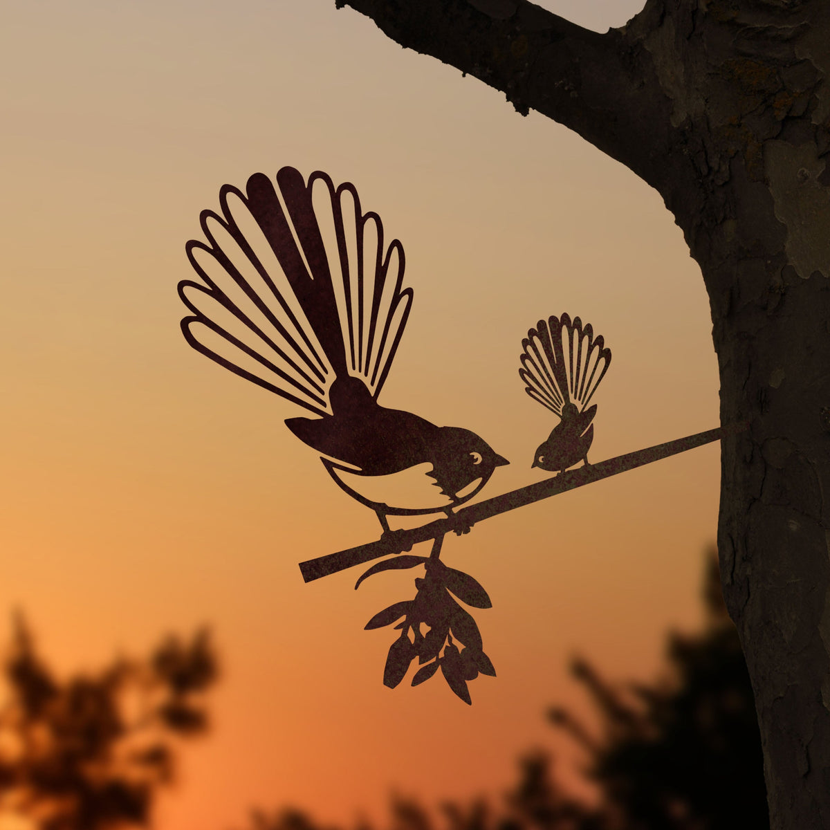 Fantail and chick steel silhouette on a branch beside a tree at sunset.
