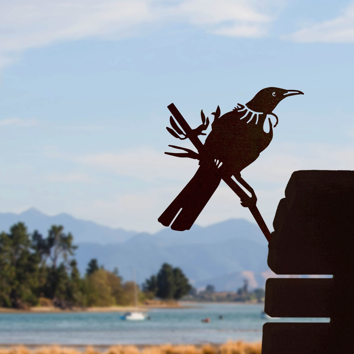Tūī steel silhouette mounted beside a timber post with a coastal view.